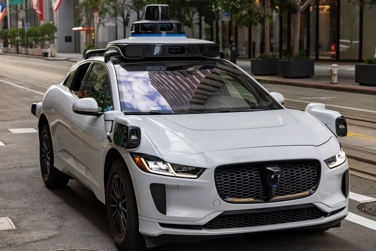 A white autonomous vehicle navigating a city street, reflecting urban architecture in daylight.